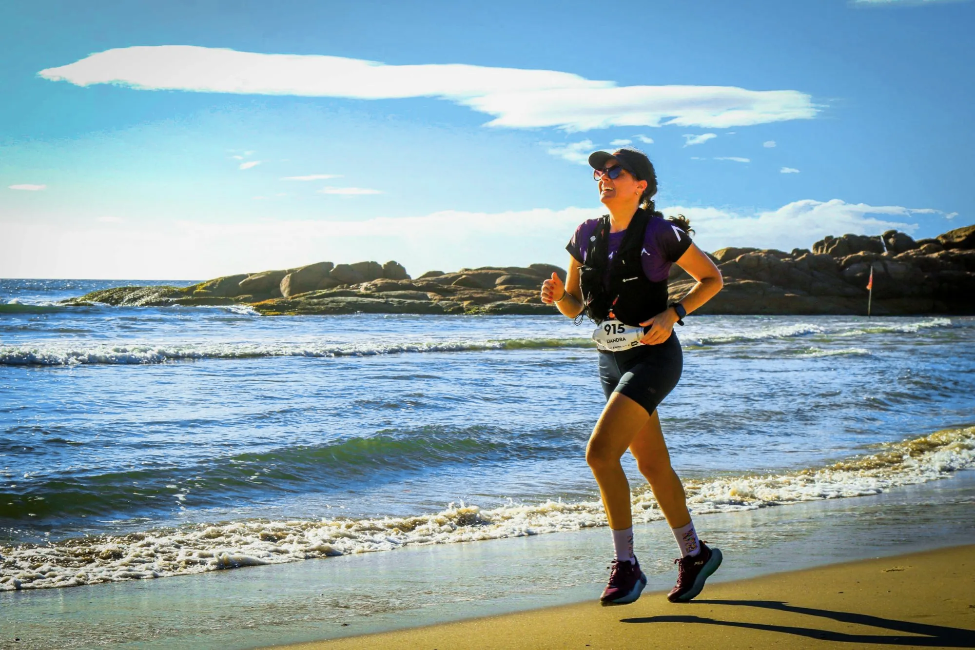 Atleta corre na areia, à beira do mar, com o costão de pedras ao fundo